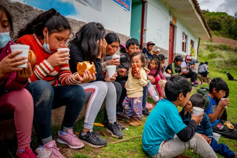 Niños y jóvenes de la comunidad comparten pan y una bebida sentados junto al local comunal